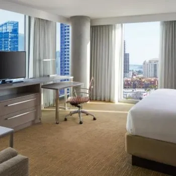 Modern hotel room with a large bed, desk, TV, armchair, and windows offering a city and water view.