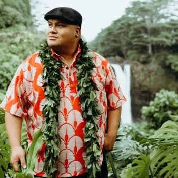 A man in a red and white patterned shirt and leafy lei stands outdoors among lush greenery, with a waterfall and trees in the background.