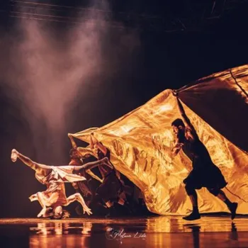 Dancers perform on stage; one balances on their head while others manipulate a large golden fabric under dramatic lighting.
