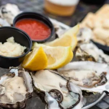 A plate of raw oysters on the half shell served with lemon wedges, cocktail sauce, horseradish, and crackers on a mosaic-style table.