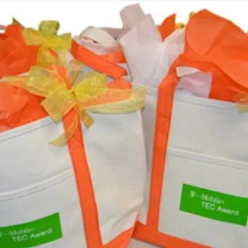 Three white tote bags with orange handles, each filled with tissue paper and decorated with a yellow ribbon and a green label reading "T-Mobile TEC Award.