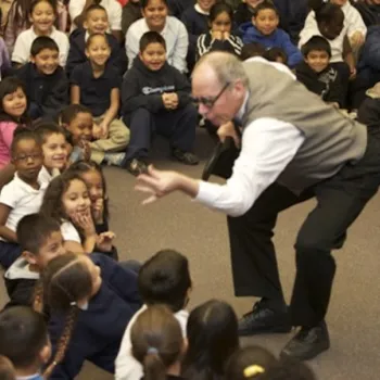 A man interacts animatedly with a large group of young children seated on the floor, who are watching and smiling attentively.