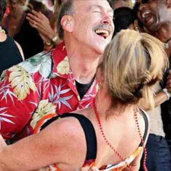 A man in a red floral shirt and a woman dance together in a crowded, festive setting, smiling and enjoying themselves among other dancing couples.