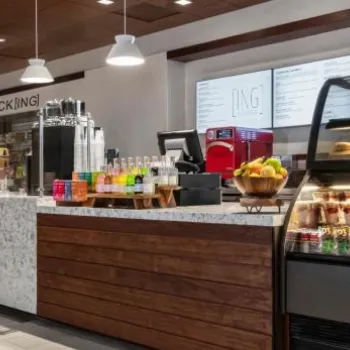 Modern self-serve café area with a counter displaying drinks, snacks, and pastries, refrigerators with bottled beverages, and menu boards on the wall.