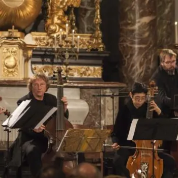 Five musicians perform classical music with string instruments in an ornate hall, seated in front of an audience and elaborate gold and marble decor.