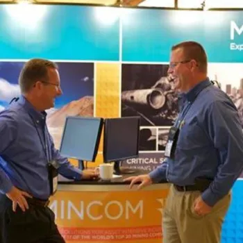 Two men stand and talk at a Mincom exhibition booth, with promotional materials and computer monitors visible in the background.