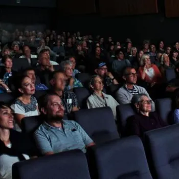 A large audience sits in a darkened movie theater, facing the screen, with most seats occupied and people watching attentively.