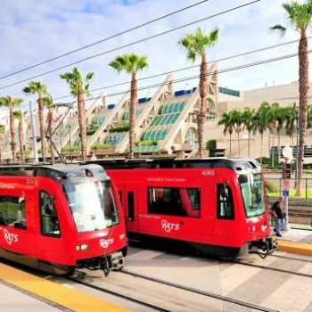 Two red trolley trains at a station platform with palm trees and a modern building in the background, under a partly cloudy sky.
