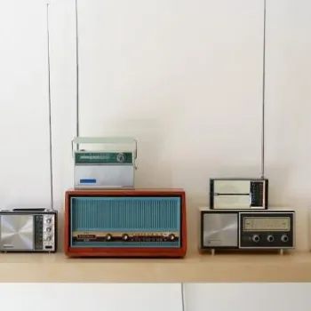 A collection of seven vintage radios with antennas displayed on a floating wooden shelf against a plain white wall.
