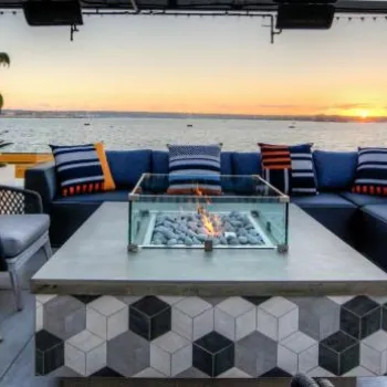 Outdoor seating area with a fire pit, modern furniture, and striped cushions overlooks the water at sunset, with boats and a tall ship visible in the background.