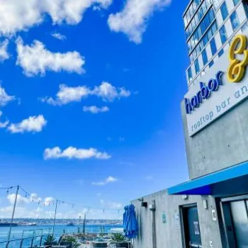 Outdoor patio area of a rooftop bar and lounge overlooks a body of water under a blue sky with scattered clouds; string lights hang above the seating area.