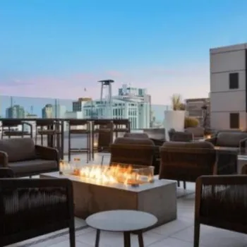 Outdoor rooftop lounge with modern seating, a fire pit in the center, tall tables, city buildings in the background, and a clear evening sky.