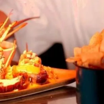 A person serves a plate of grilled lobster garnished with lime, accompanied by a small bowl of sauce and a side of bread or chips in a separate container.