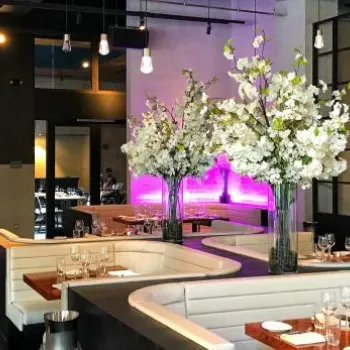 Modern restaurant interior with white booths, wooden tables, large flower arrangements, and purple accent lighting on brick walls.