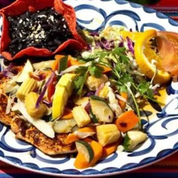A colorful plate of grilled chicken with mixed vegetables, salad, sliced tomato, and a small red bowl of black beans on a vibrant striped tablecloth.