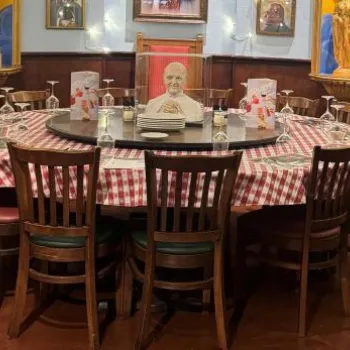 A round dining table with a red and white checkered tablecloth is surrounded by wooden chairs in a restaurant, with framed portraits and gold columns in the background.