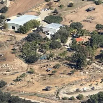 Aerial view of a rural property with multiple buildings, winding dirt paths, fenced areas, scattered trees, and patches of cultivated land.