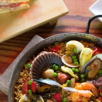 A pan of seafood paella with shrimp, mussels, and vegetables, a slice of savory pie on a wooden board, and a plate with salad, grilled bread, and sauce.
