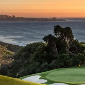A coastal golf course green with surrounding sand bunkers overlooks the ocean at sunset, with distant hills and cityscape in the background.