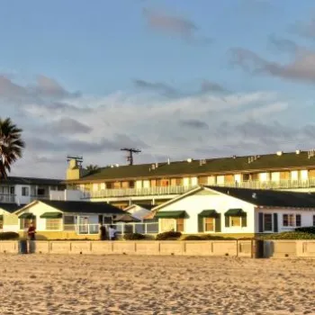 A row of beach cottages and a three-story hotel stand along a sandy beach under a partly cloudy sky, with palm trees and parked cars visible.