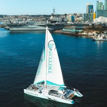 A white catamaran with "TRITON" on its sail is sailing in a bay near a city skyline, with a large navy ship and buildings visible in the background.