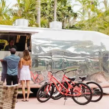 Two people stand at the window of a shiny silver food truck surrounded by palm trees, with two red bicycles parked nearby and outdoor seating in the foreground.