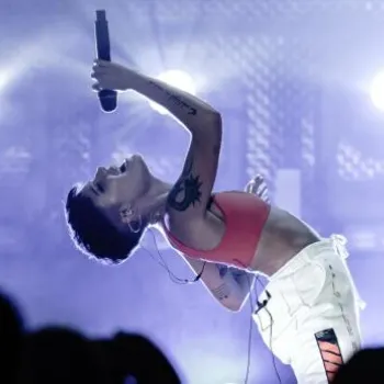 A performer in a red top and white pants sings on stage, leaning back dramatically with a microphone, in front of bright stage lights and an audience.