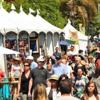 A large crowd of people walks through an outdoor art festival with white tents displaying artwork under sunny weather.