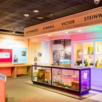 A brightly lit museum exhibit displays vintage musical instruments, including guitars and drums, with signage for brands like Steinway and Wurlitzer above a replica music store counter.