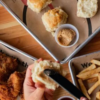 Overhead view of trays with fried chicken, biscuits, fries, dipping sauces, and cups of soda on a wooden table. A hand spreads butter on a biscuit.