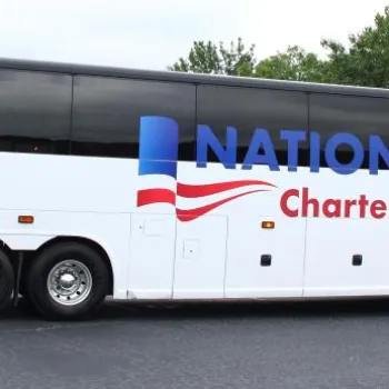 A large white charter bus with "National Charter Bus" and a blue and red logo displayed on the side, parked on an asphalt surface near trees and grass.
