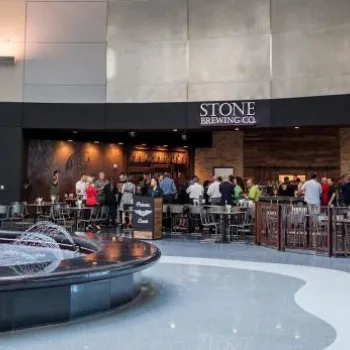 People gather at the Stone Brewing Co. restaurant inside an airport terminal, with a decorative fountain in the foreground and shops visible to the side.
