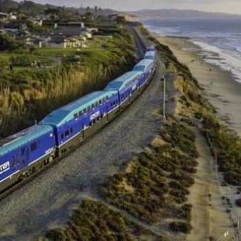A blue passenger train travels along tracks beside a sandy beach and ocean, with a grassy park and houses on the left and hills in the distance.