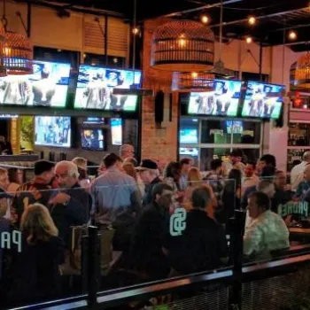 A crowded sports bar with people seated at tables, several TVs showing sports, and neon signs visible in the background.