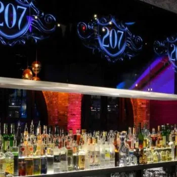Modern bar with various bottles of liquor on shelves, illuminated by colorful lights, with "207" sign above and abstract art displayed on a screen in the background.