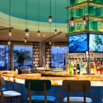 Modern bar with illuminated counter, hanging lights, multiple screens, and marina views through large windows in the background.