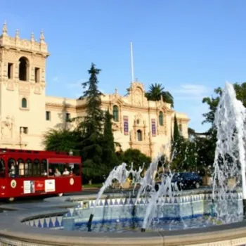 A red trolley passes by a historic Spanish-style building and a decorative fountain in a landscaped plaza on a sunny day.