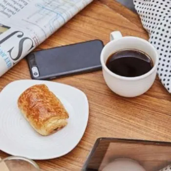 A person sits at a table with a cup of coffee, a glass of orange juice, a pastry on a plate, a newspaper, a phone, and a tablet.