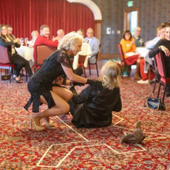 Two women are on the floor in the center of a banquet hall, one helping the other up, while guests seated at round tables watch and react.