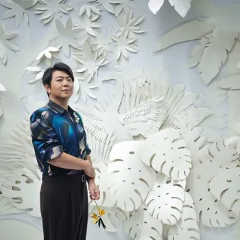 A person stands in front of a white, layered paper backdrop featuring plants and a bird, holding a yellow flower and wearing a blue patterned shirt.