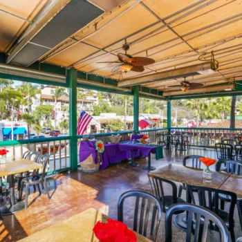 Outdoor restaurant patio with empty tables and chairs, ceiling fans, and potted red flowers; view of buildings, trees, and American flag in the background.