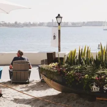 People sit in lounge chairs on a sandy beach overlooking the water, with a Seaport Village planter and an umbrella in the foreground.