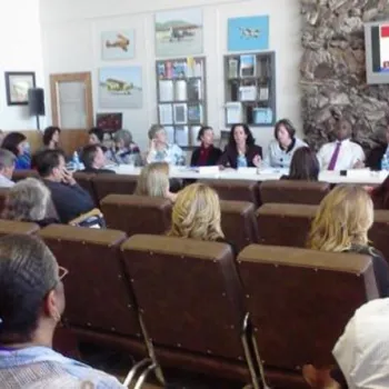 A group of people attend a panel discussion in a conference room with a presentation projected on a screen at the front.