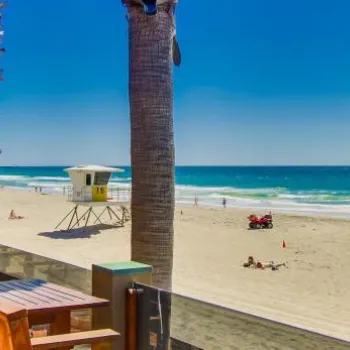 Wooden tables and chairs overlook a sandy beach with palm trees, a lifeguard tower, and people enjoying a sunny day by the ocean.