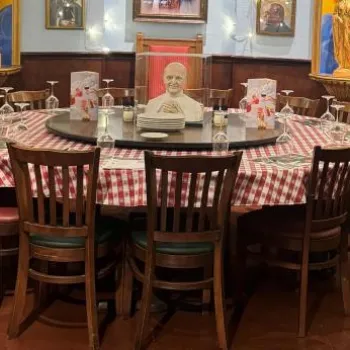 A large round table with a red and white checkered tablecloth is set for a meal, surrounded by wooden chairs in a restaurant with framed portraits and gold columns.