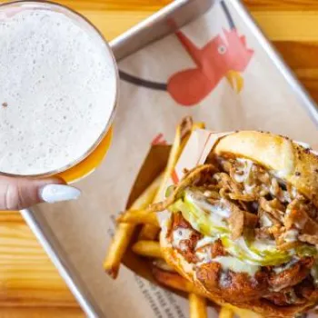 A person holds a glass of beer in one hand and a loaded burger in the other, with fries on a tray on a wooden table.