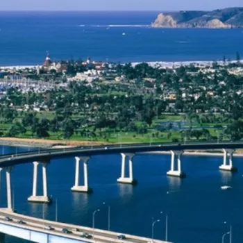 A curved bridge crosses over blue water with cars driving on it, leading to a coastal city with greenery, buildings, and hills in the background.