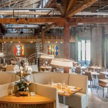 Spacious modern restaurant interior with wooden beams, high ceilings, contemporary artwork, and tables set for dining. Natural light enters through large windows.
