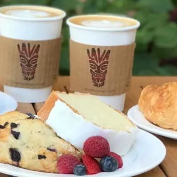 Two cups of coffee, a chocolate chip muffin, a croissant, a slice of pound cake with berries, and a blueberry scone are arranged on white plates on a wooden table outdoors.