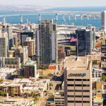 Aerial view of downtown San Diego featuring high-rise buildings, the bay, and the Coronado Bridge in the background on a clear day.